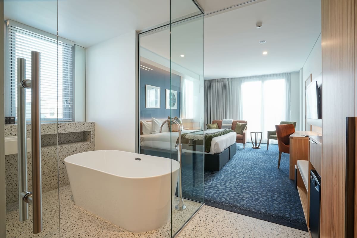 Ein modernes Hotelzimmer mit einem gläsernen Badezimmer, einer großzügigen Badewanne, einem Bett mit blauer Kopfteil, einem Schreibtisch und einem Stuhl.