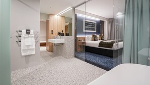 A modern hotel room with a glass partition, a bed, a bedside table, a built-in TV, and a bathroom with a sink and towel rack. - The Langlands Hotel (Invercargill)
