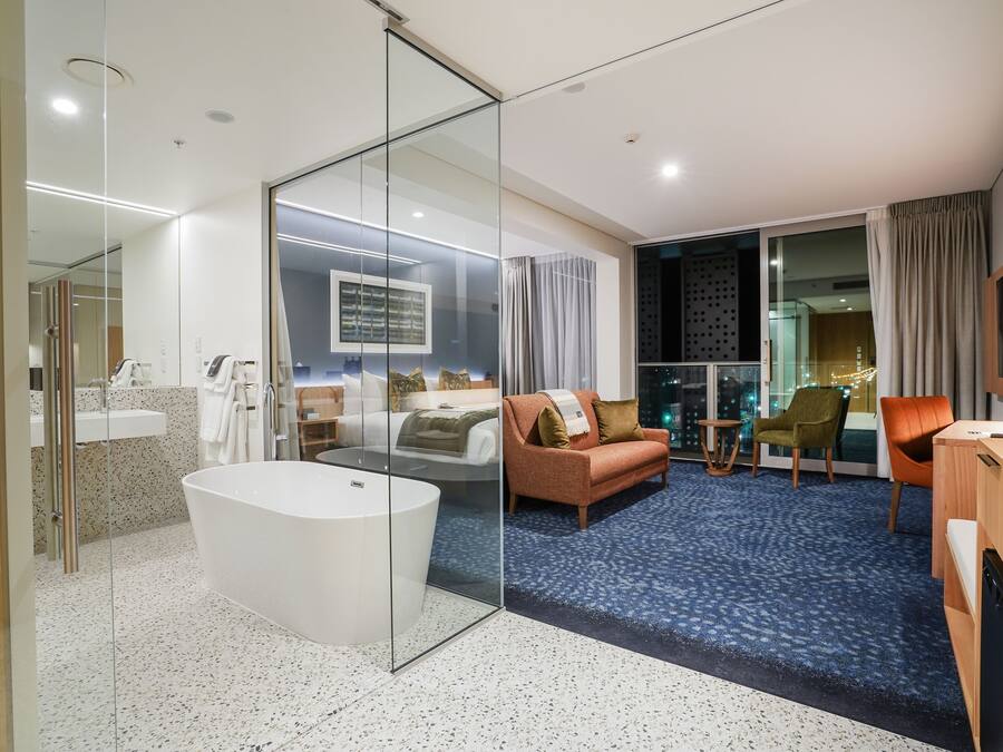 A modern hotel room with a glass-enclosed shower, a large bathtub, a sofa, a coffee table, and a view of the city at night.
