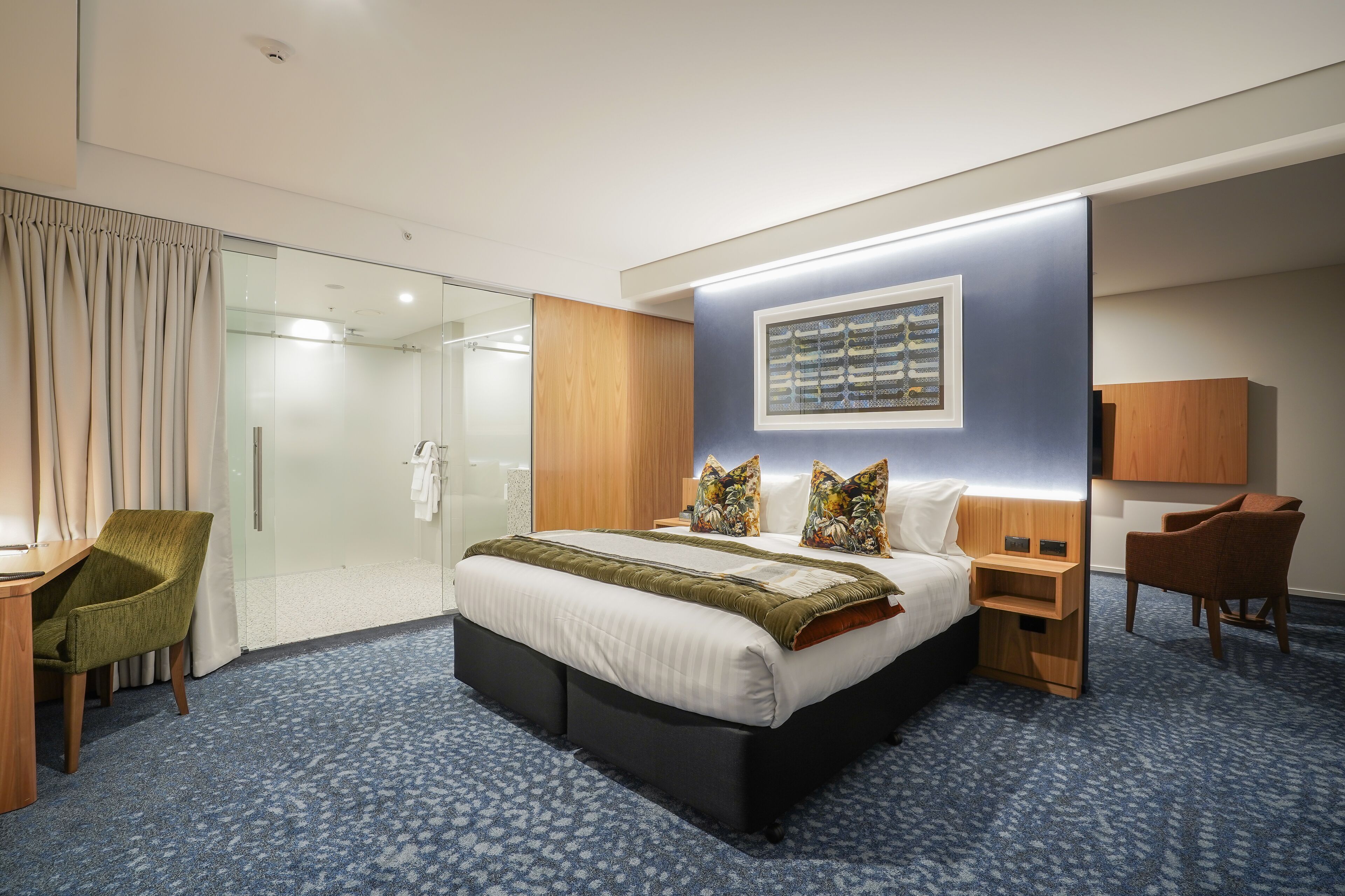 A modern hotel room with a large bed, a desk, a chair, a shower area, and a mounted artwork.