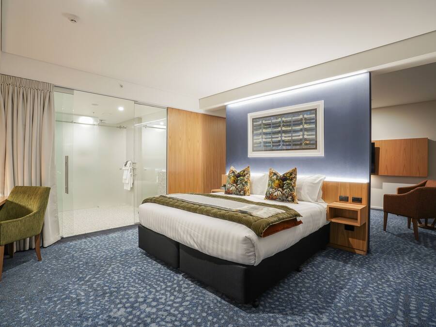 A modern hotel room with a large bed, a desk, a chair, a shower area, and a mounted artwork.