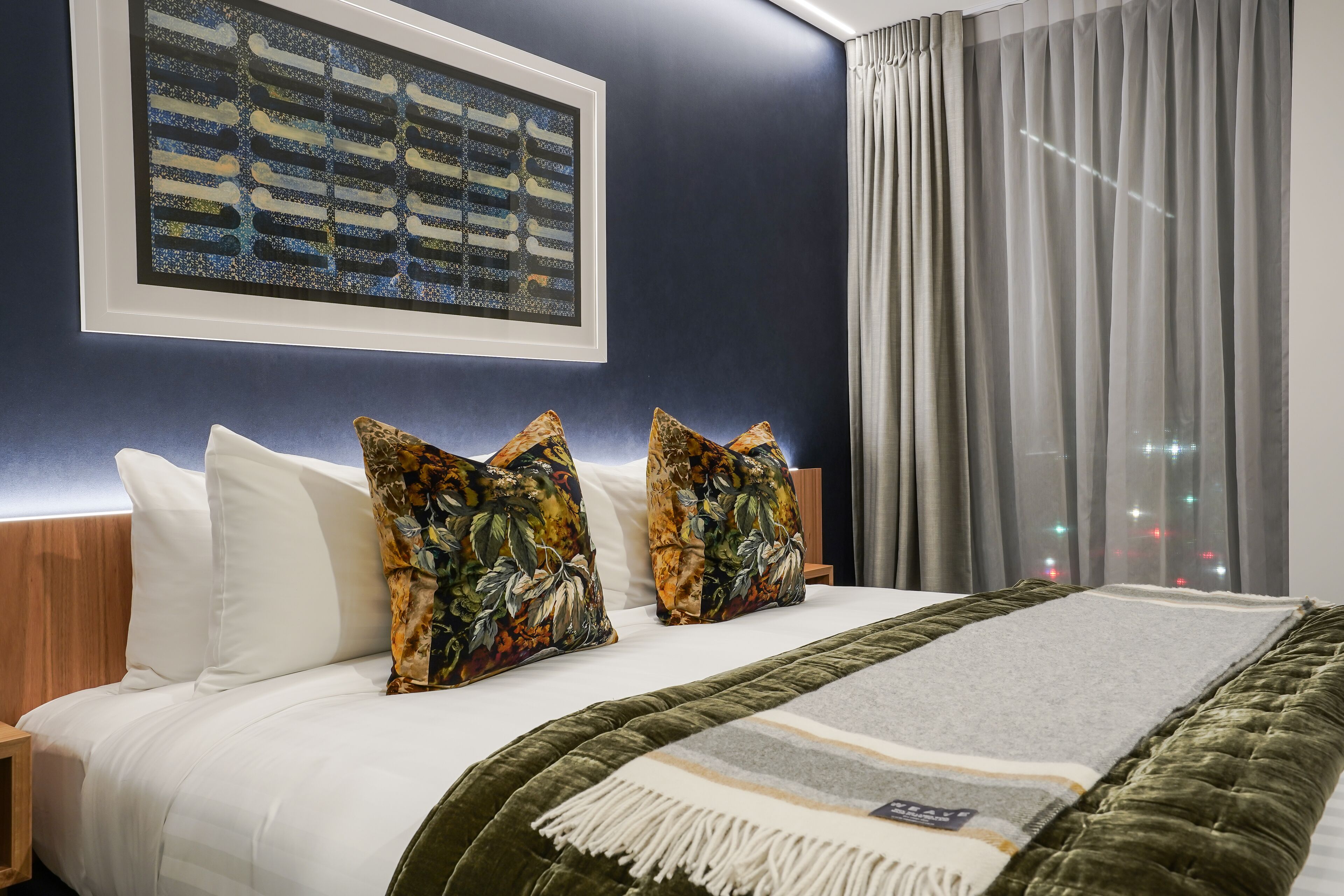 A hotel room with a large bed, two decorative pillows, a framed artwork on the wall, and a view of the city through the window.
