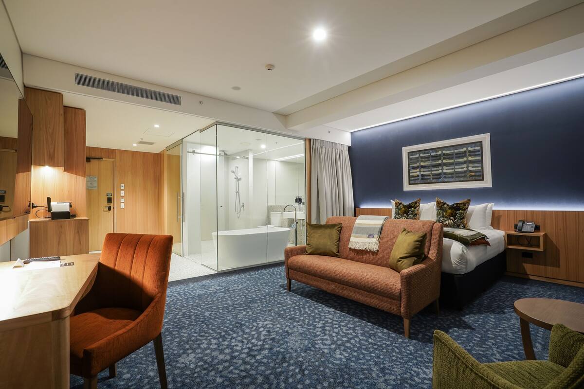 Ein Hotelzimmer mit Holzschreibtisch, einer Couch, einem Bett und einem durch eine Glasabtrennung einsehbarer Badezimmer.