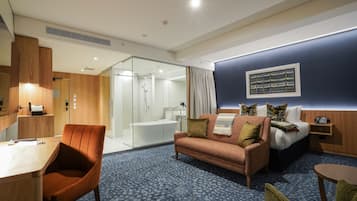 Habitación de hotel con un escritorio de madera, un sofá, una cama y un baño visible a través de un panel de vidrio.