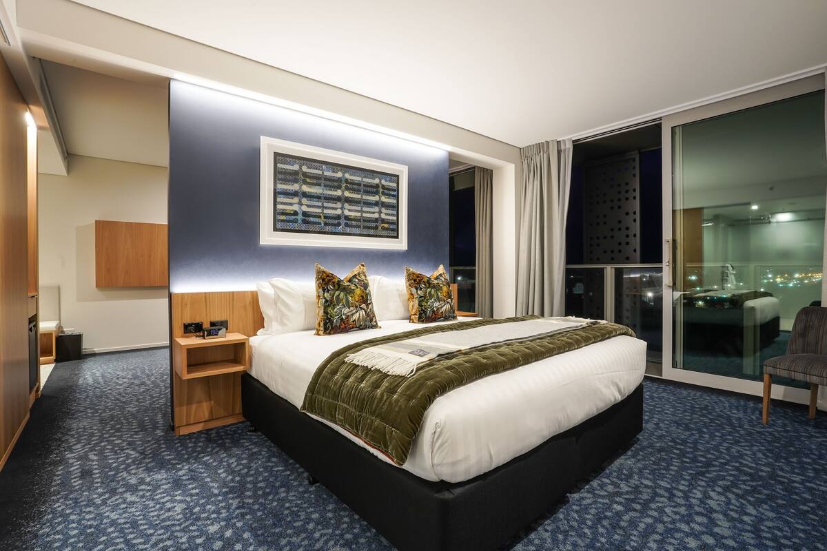 Ein Hotelzimmer mit einem großen Bett, einem Nachttisch, einer Holzablage, einem Fenster mit Vorhängen und Blick auf die nächtliche Stadt.