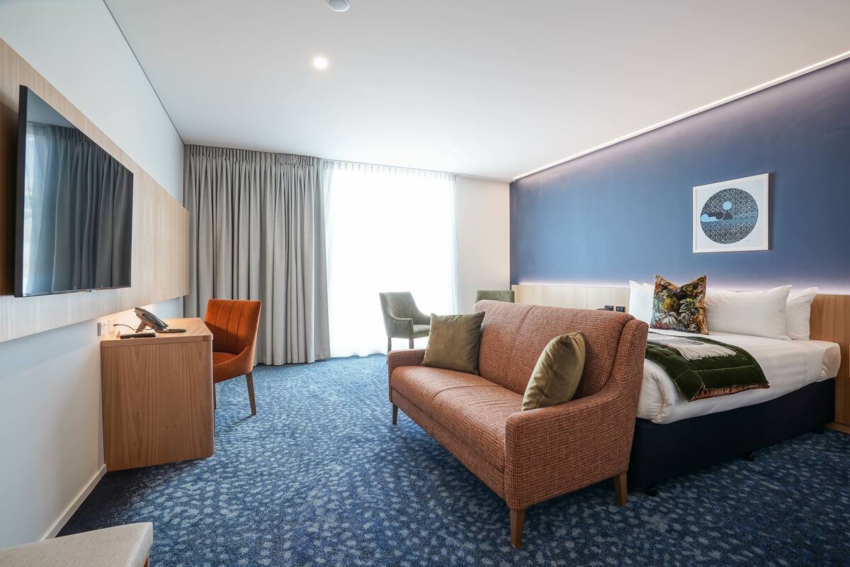 Ein Hotelzimmer mit einem großen Bett, einer Sitzecke, einem Schreibtisch und einem Sessel. Ein Fenster mit Vorhängen und ein an der Wand befestigter Fernseher sind ebenfalls vorhanden.