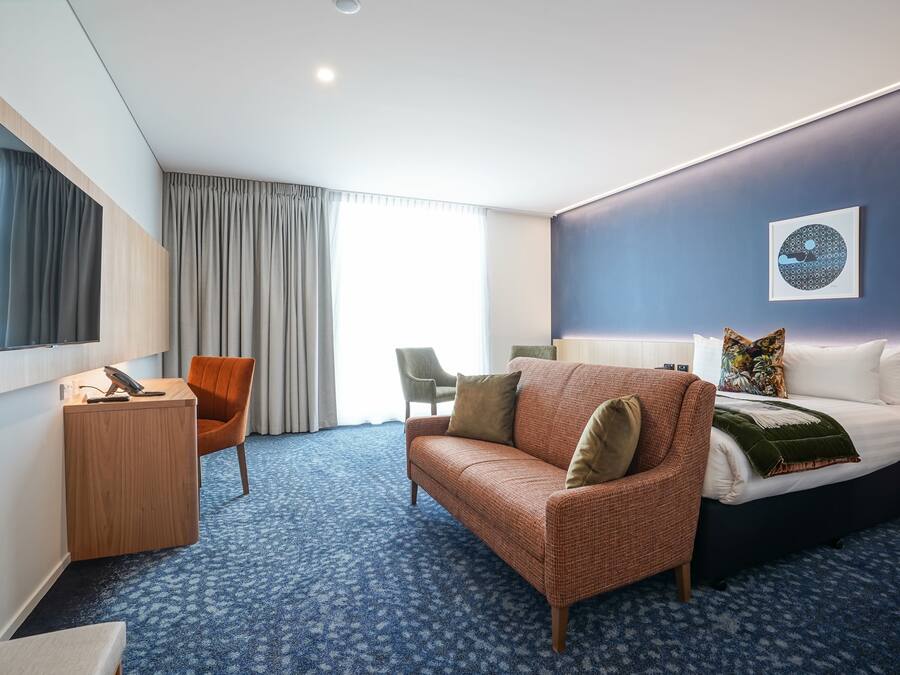 A hotel room with a large bed, a sofa, a desk, and a chair. There is a window with curtains and a wall-mounted TV.