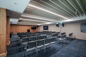 Un salón de conferencias con filas de sillas, un atril, una pantalla y un panel de pared de madera.