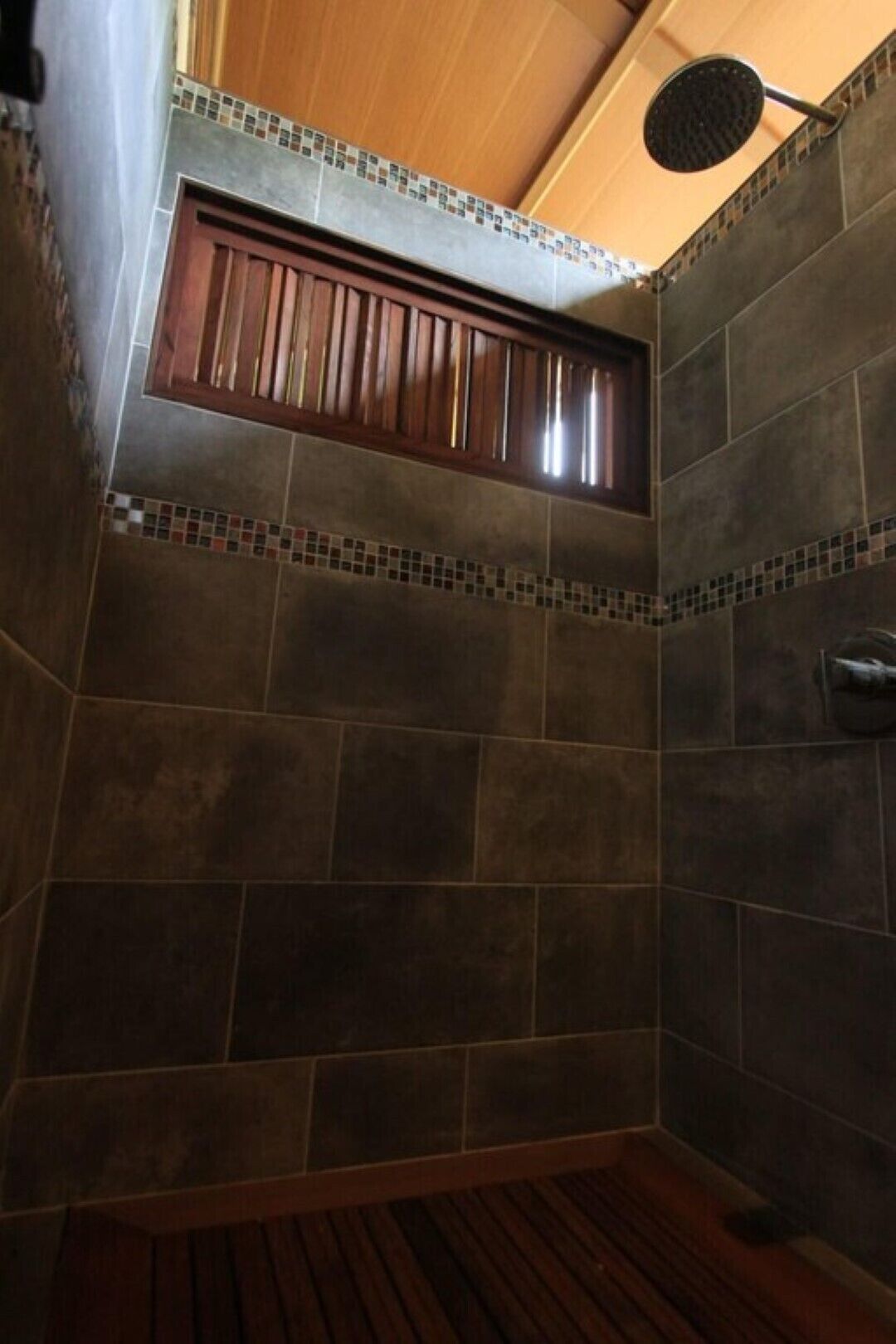Una ducha con azulejos oscuros, un banco de madera y un cabezal de ducha.