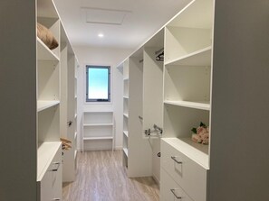 A narrow walk-through closet with multiple shelves and drawers, a window at the end, and a light-colored wooden floor.