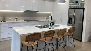 Une cuisine moderne avec des meubles blancs, un réfrigérateur en acier inoxydable, un îlot central intégré avec des tabourets, un éclairage suspendu et une machine à café.