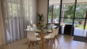 Un coin repas avec une table en verre, des chaises blanches et une vue sur la verdure grâce à de grandes fenêtres.