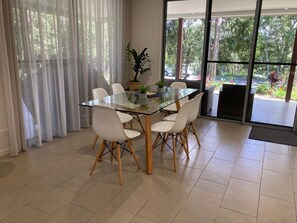 A dining area with a glass table, white chairs, and a view of greenery through large windows.