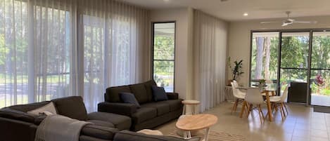 A modern living room with a sectional sofa, a dining table with chairs, and large windows with sheer curtains.