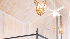 Una cama con dosel, cubierta con una colcha acolchada y almohadas, un ventilador de techo e lámparas colgantes en una habitación con paredes de madera y una puerta visible al fondo.