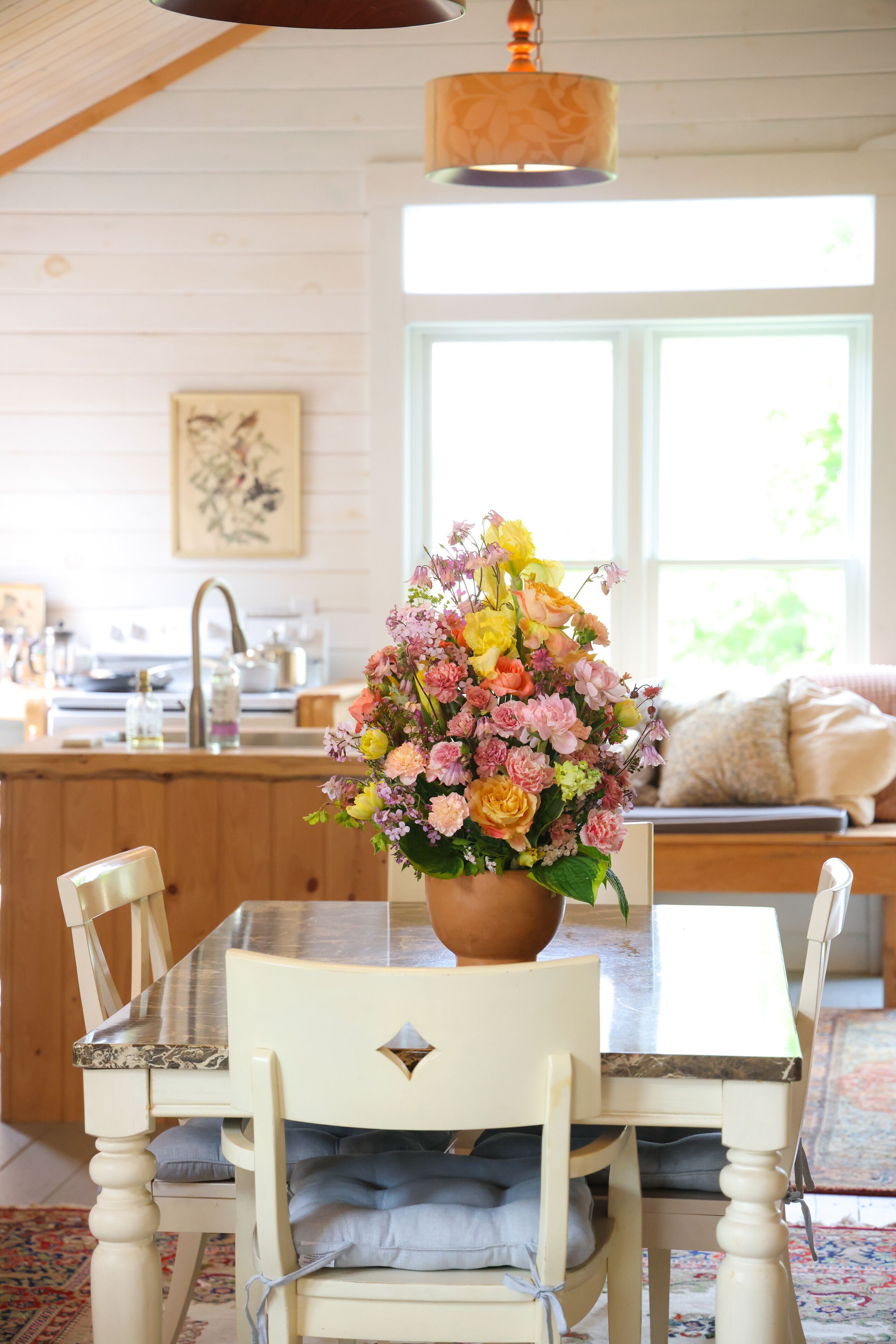 A dining area with a table, chairs, a vase of flowers, a kitchen in the background, and a window with a view.