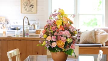 A dining area with a table, chairs, a vase of flowers, a kitchen in the background, and a window with a view.