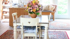Un comedor con una mesa de madera, sillas blancas, un centro de mesa floral y una lámpara colgante.