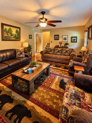 A living room with a brown leather sofa, a wooden coffee table, patterned rugs, and a ceiling fan. - Cozy Cabin Retreat Sleeps 12 w/ HotTub, Wi-Fi, FirePit minutes from Dollywood!! (Sevierville)
