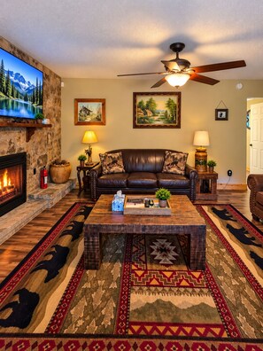A cozy living room with a fireplace, a leather sofa, a coffee table, and a television mounted on a stone wall. - Cozy Cabin Retreat Sleeps 12 w/ HotTub, Wi-Fi, FirePit minutes from Dollywood!! (Sevierville)