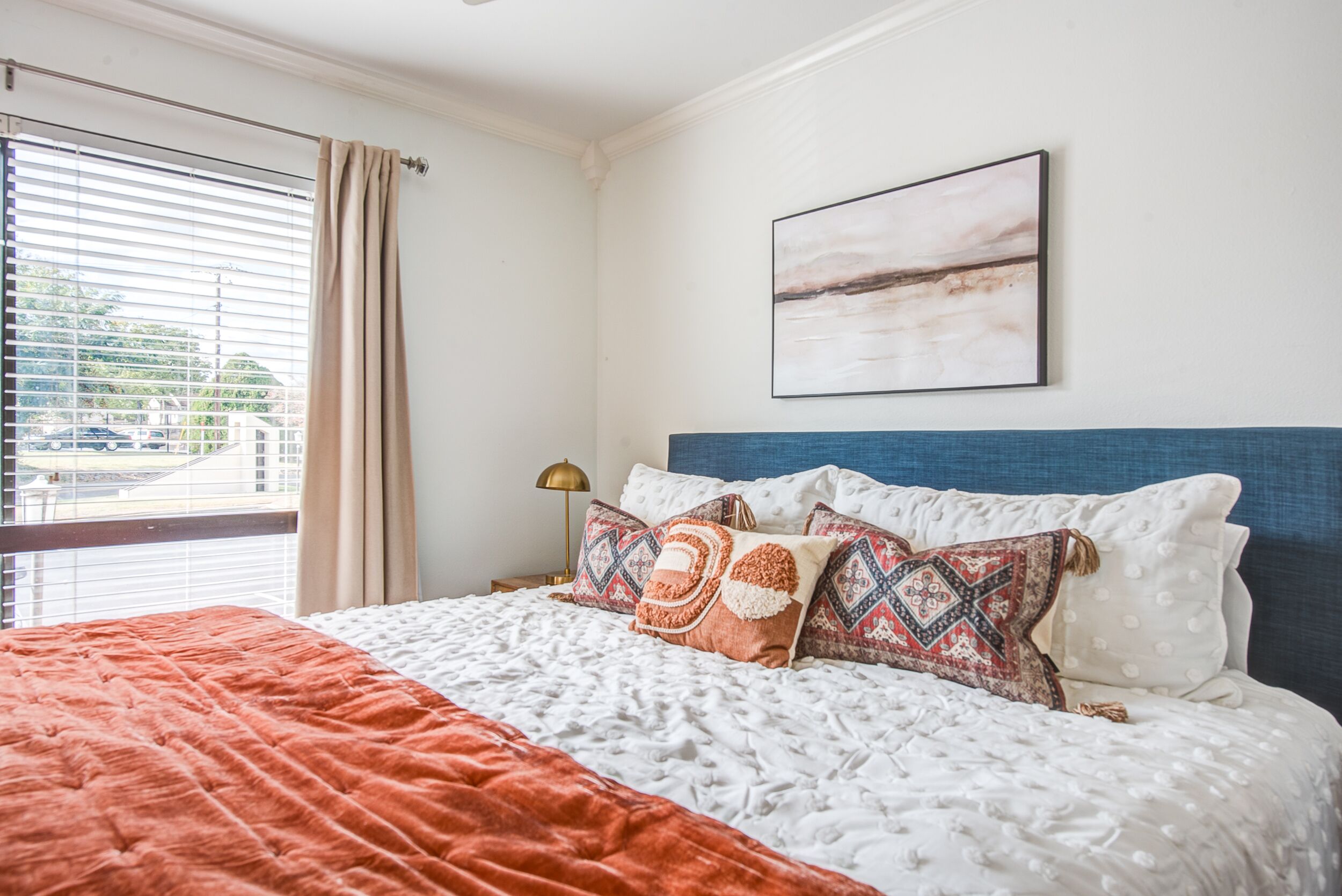 A bedroom with a large bed, a window with blinds, a wall-mounted artwork, and a nightstand with a lamp.