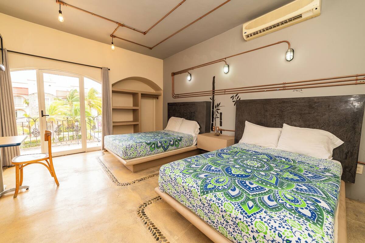 Une chambre d’hôtel avec deux lits, un bureau et une chaise. Une grande fenêtre offre une vue sur les palmiers et les immeubles.