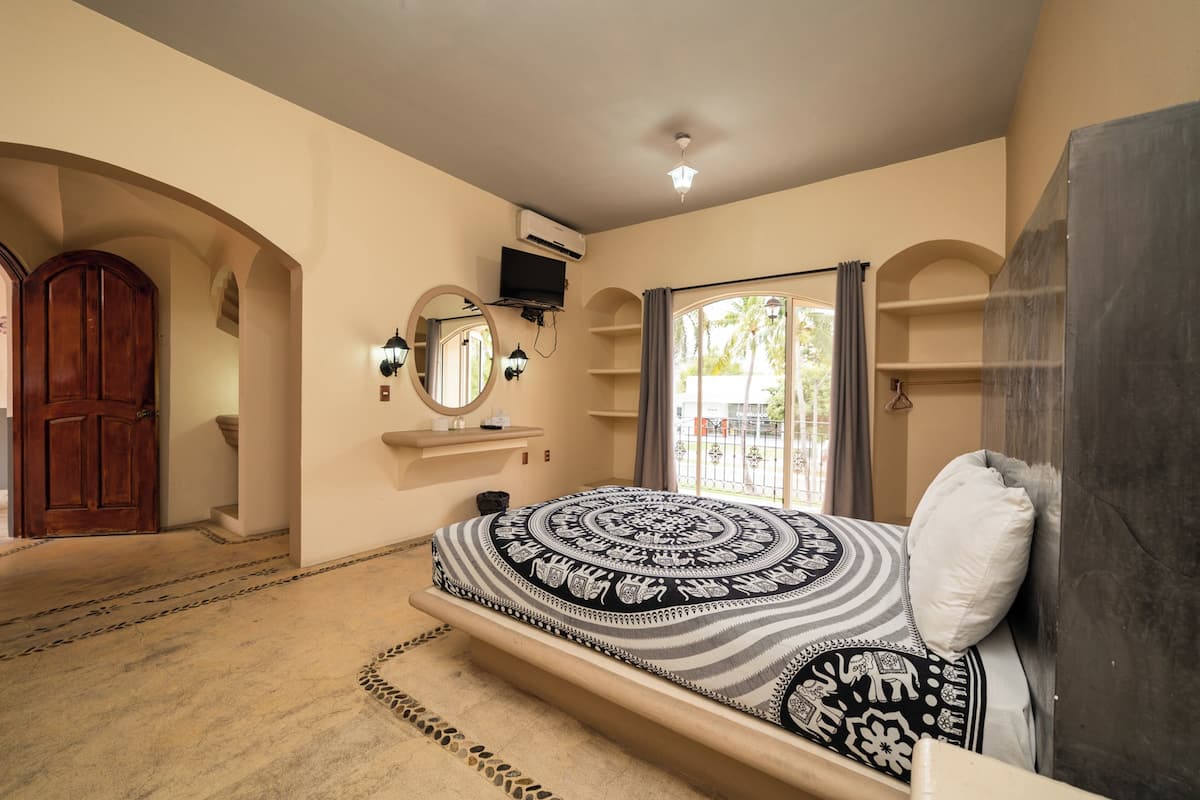 Une chambre à coucher avec un lit, une porte en bois, un appareil de climatisation, un miroir et une fenêtre avec des rideaux.