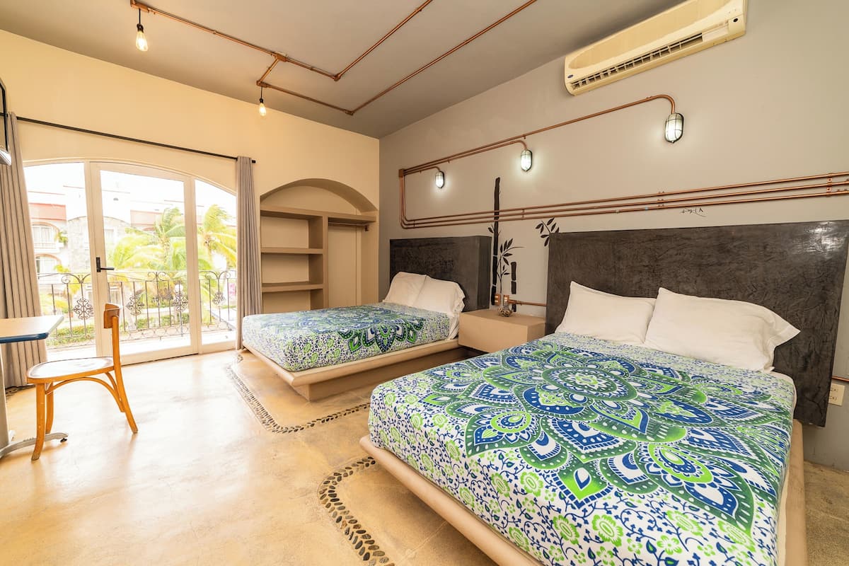 Une chambre d’hôtel avec deux lits, un bureau et une chaise. Une grande fenêtre offre une vue sur les palmiers et les immeubles.