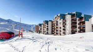 A snow-covered ski resort with modern multi-story buildings, ski lifts, and a clear blue sky.