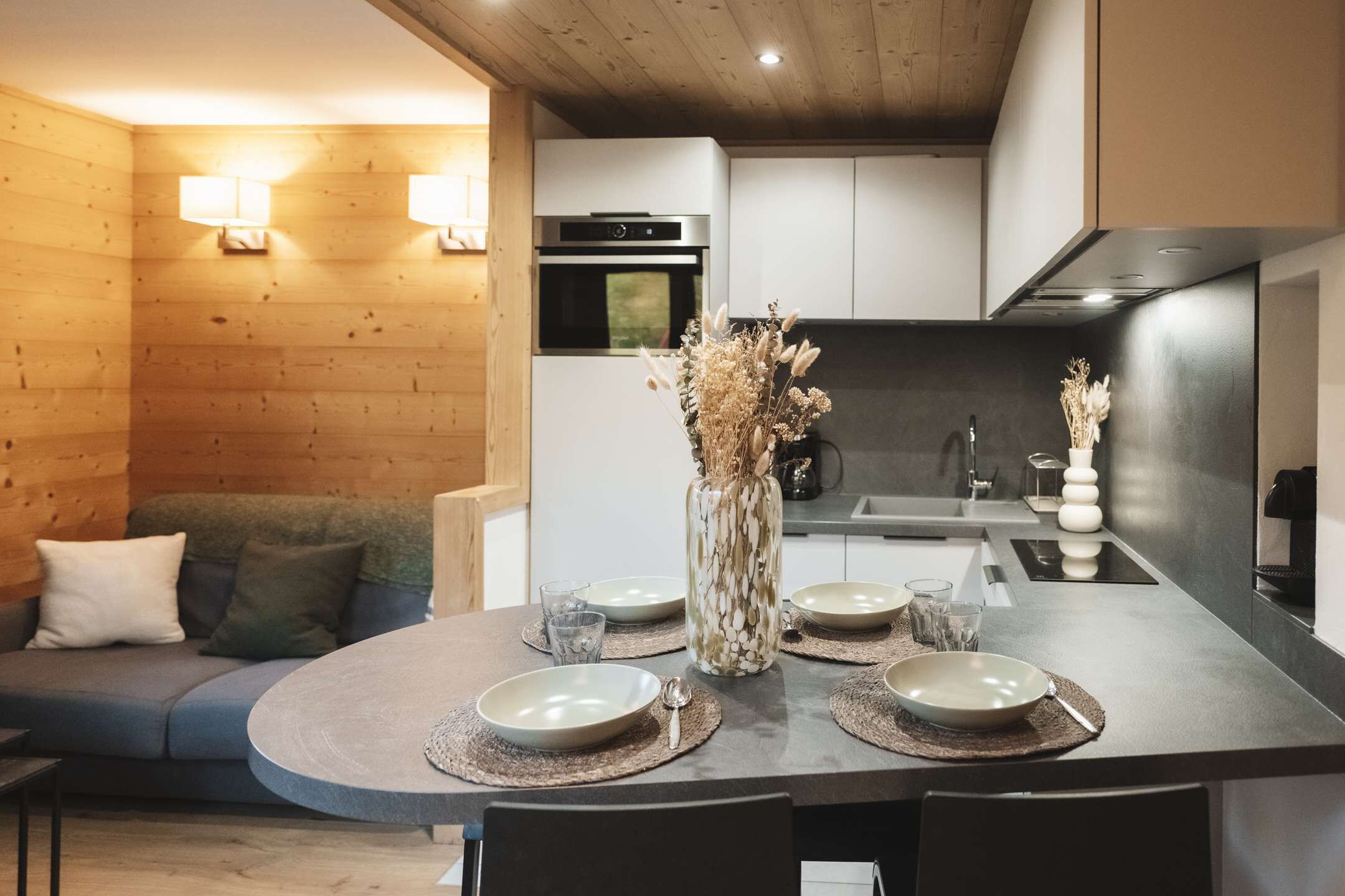 A modern kitchen with a dining area, wooden walls, and a central island with a vase of dried flowers.