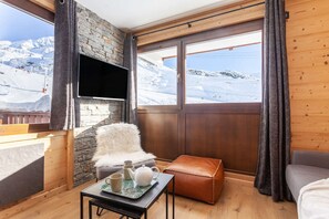 A cozy room with a stone wall, a flat-screen TV, a cushioned chair, a small table with a teapot and cups, and a view of a snowy mountain landscape.