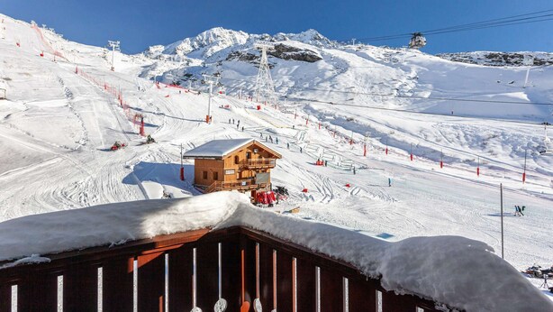 A wooden cabin with a snow-covered roof, surrounded by a ski slope with ski lifts and red safety barriers.