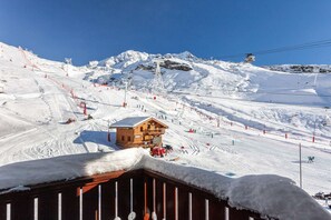 Een houten chalet met een sneeuwbedekte dak, omringd door een piste met skiliften en rode vangrails.
