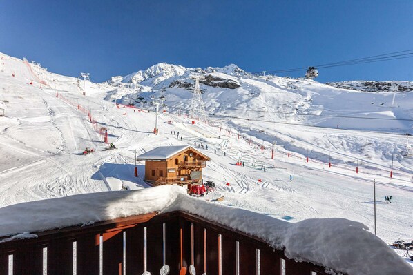 Un chalet en bois, dont le toit est recouvert de neige, entouré d’une piste de ski avec des remontées mécaniques et des barrières de sécurité rouges.