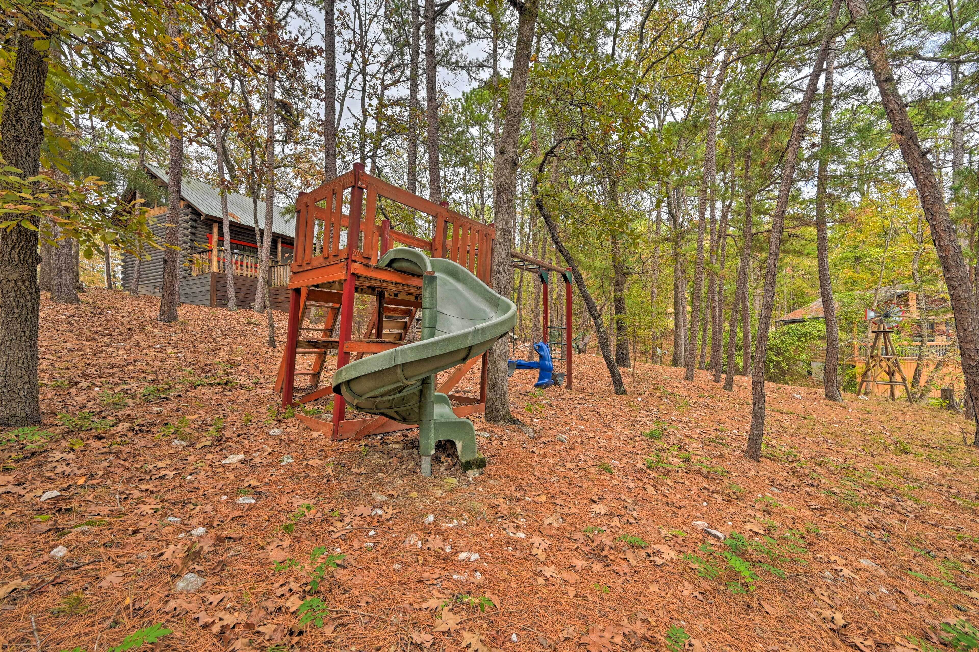 Ein Spielplatz mit Rutsche und Schaukeln, umgeben von Bäumen und einem Häuschen im Hintergrund.
