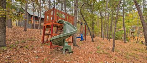 Ein Spielplatz mit Rutsche und Schaukeln, umgeben von Bäumen und einem Häuschen im Hintergrund.