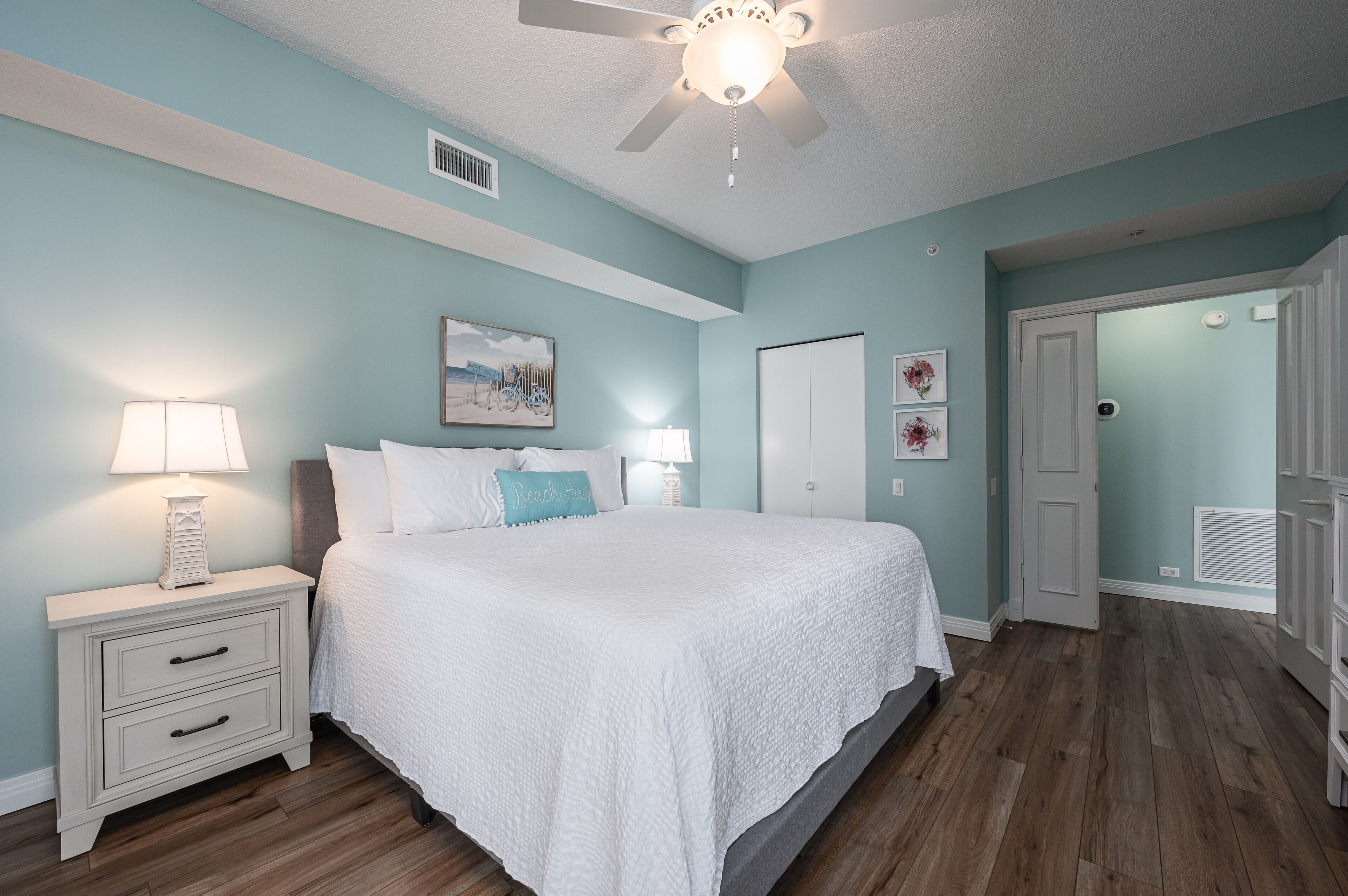 A bedroom with a bed, nightstands, a ceiling fan, and a door leading to another room.