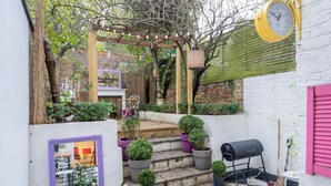 A small garden with steps leading to a wooden pergola, a barbecue grill, and a clock on the wall. - Stylish flat with private yard in Shepherd’s Bush (London)