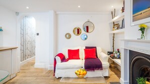 A modern living room with a white sofa, red cushions, a glass coffee table with fruit, and a fireplace. - Stylish flat with private yard in Shepherd’s Bush (London)