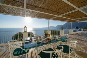 An outdoor dining area with a table set for a meal, overlooking a scenic view of the sea and mountains.