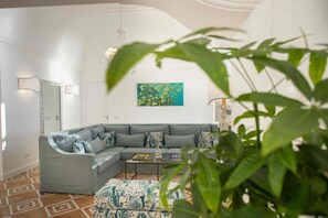 A modern living room with a grey sectional sofa, patterned ottoman, and a coffee table. A painting is on the wall, and there is a plant in the foreground.
