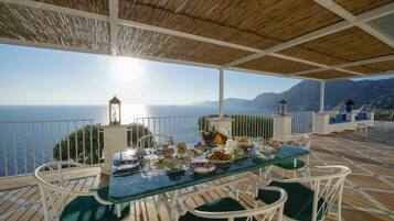 An outdoor dining area with a table set for a meal, overlooking a scenic view of the sea and mountains.