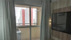 Un salón con televisor grande, un balcón con vista a edificios altos y una puerta corrediza de vidrio.