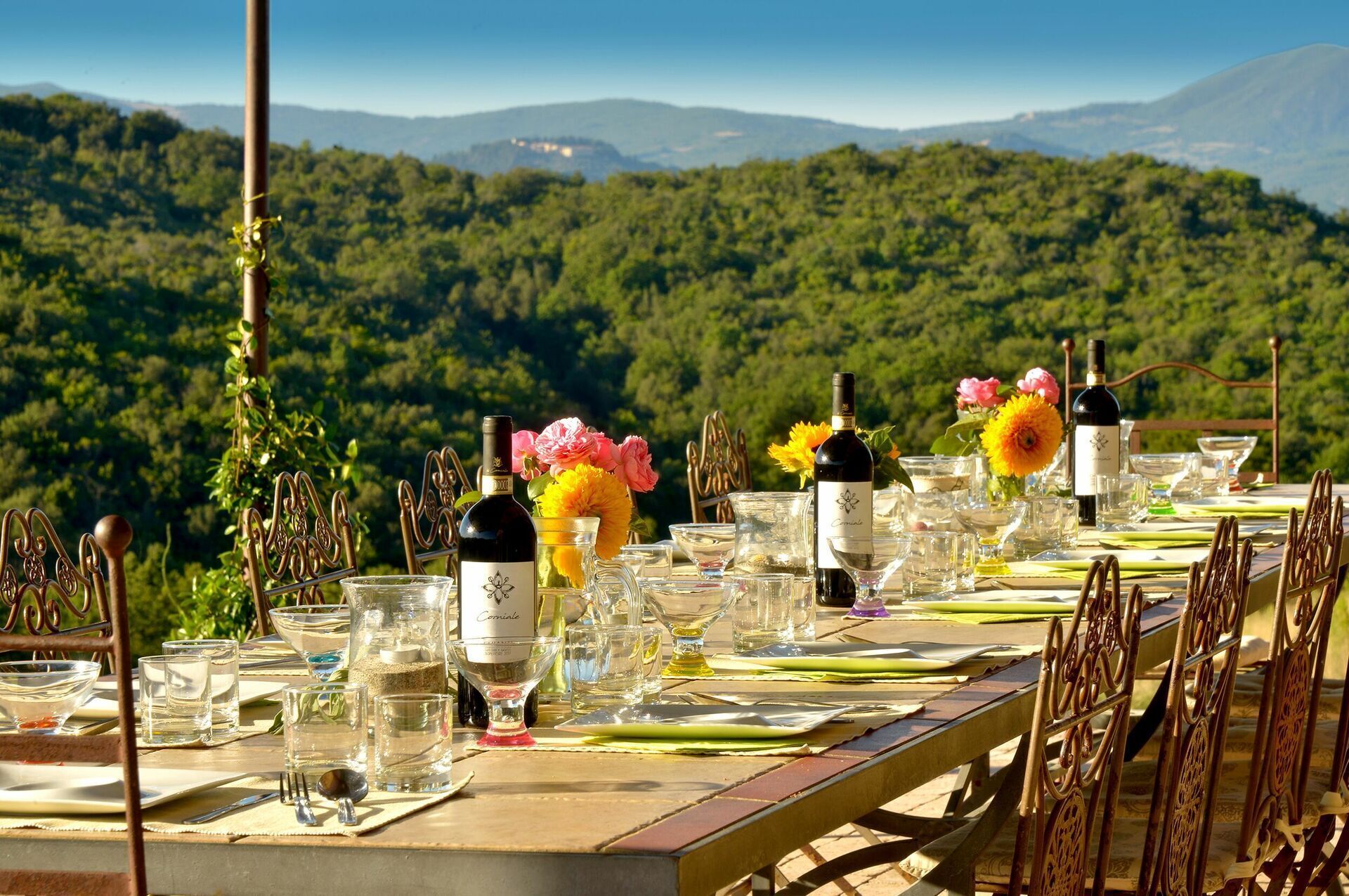 A table set for a meal with wine glasses, bottles, and cutlery, overlooking a forested mountain range.