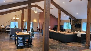 Un amplio salón con techos altos, vigas de madera a la vista, chimenea, un sofá grande, una mesa de comedor con sillas y un televisor.