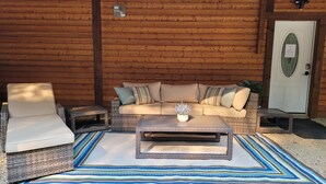 Un rincón al aire libre acogedor con un sofá acolchado, una silla y una mesa de centro, todo frente a un muro de madera.