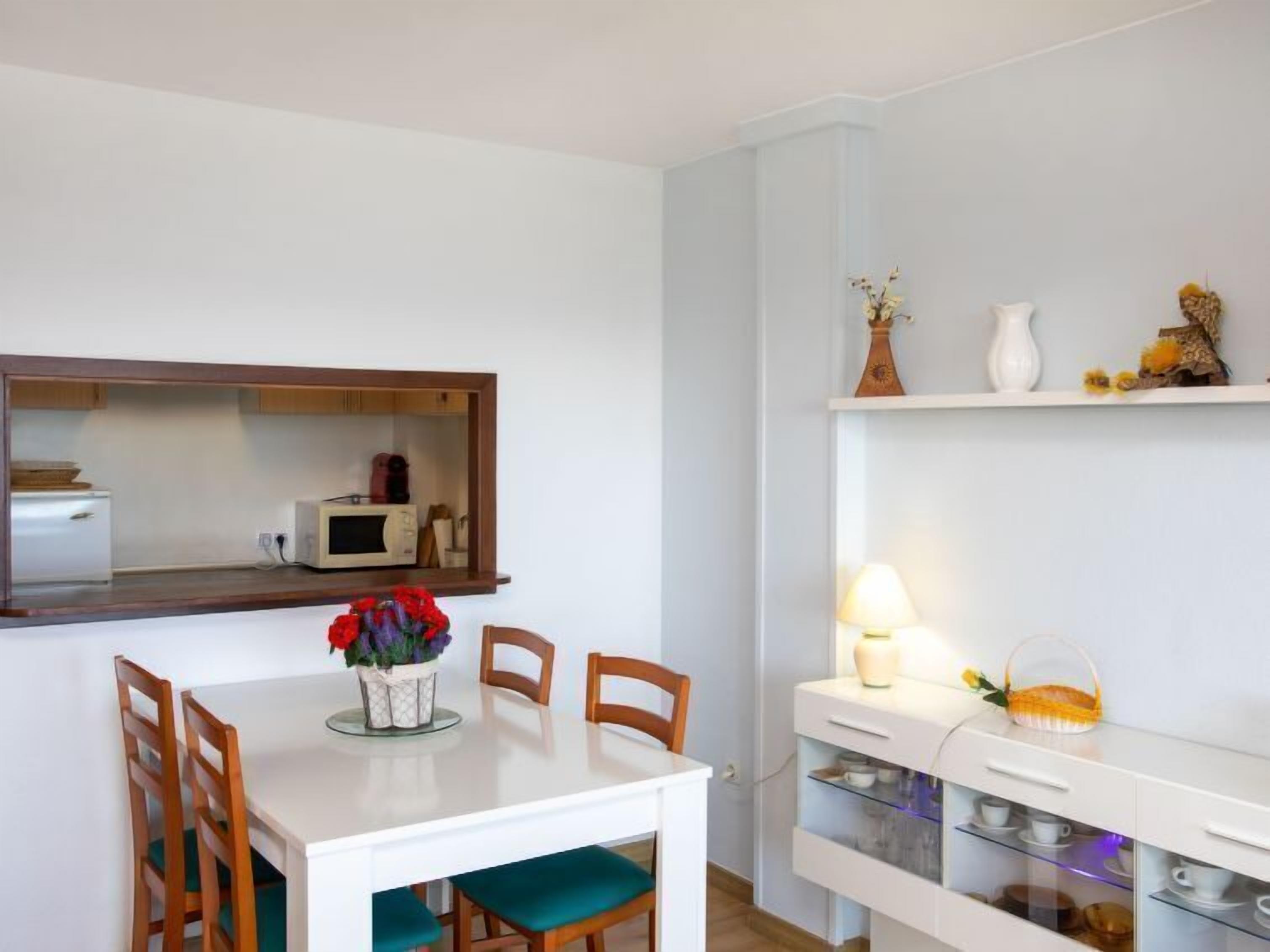 Un comedor con una mesa blanca, cuatro sillas de madera, un florero con flores rojas, y una zona de cocina con microondas y armarios.