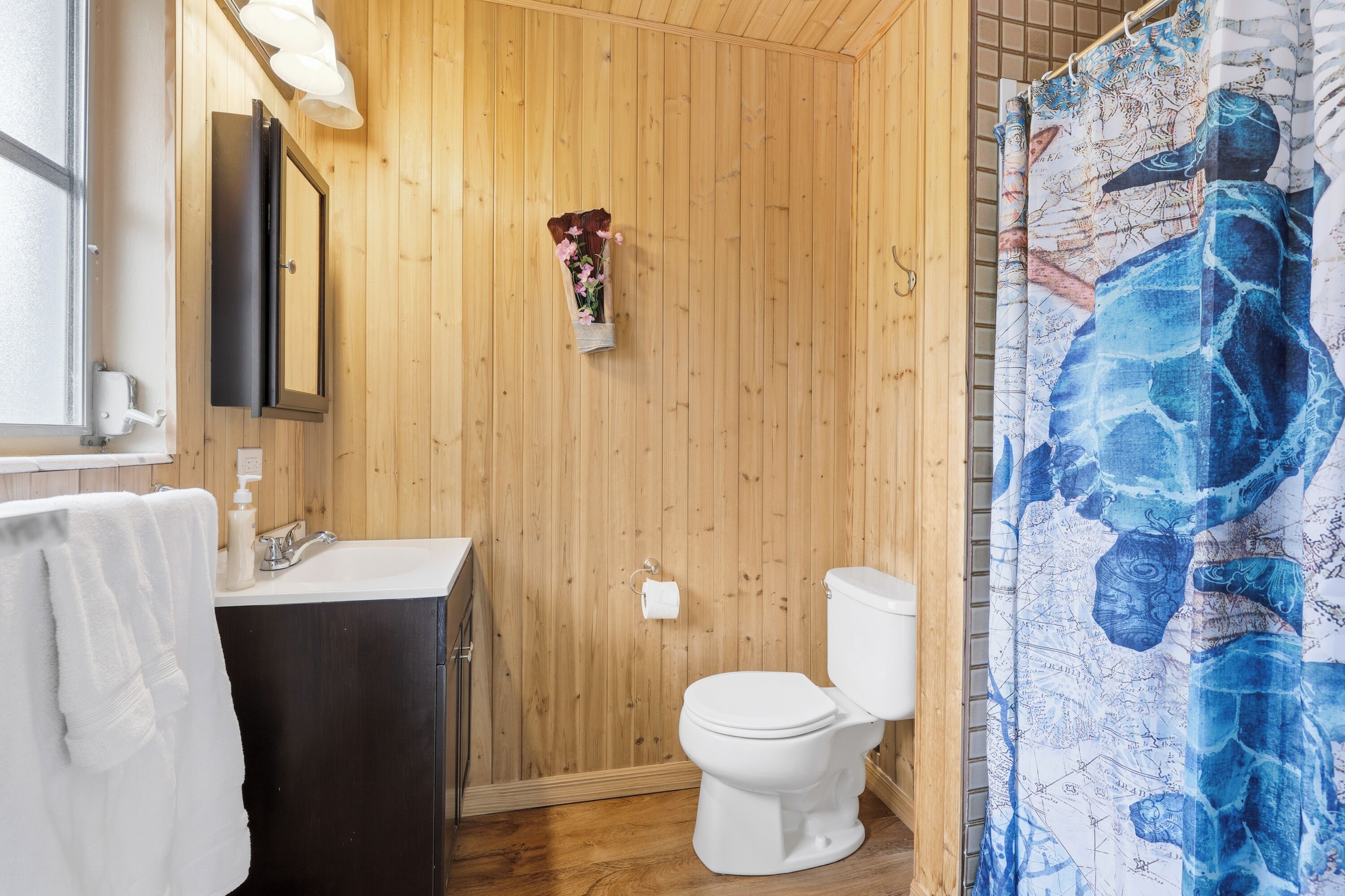 Un baño con paneles de madera, un inodoro blanco, un lavamanos con espejo y una cortina de ducha con estampado azul.