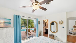 Ein Schlafzimmer mit einem großen Bett, einem Deckenventilator, Blick auf einen Pool und einem Badezimmer mit Spiegel und Handtuchhalter.