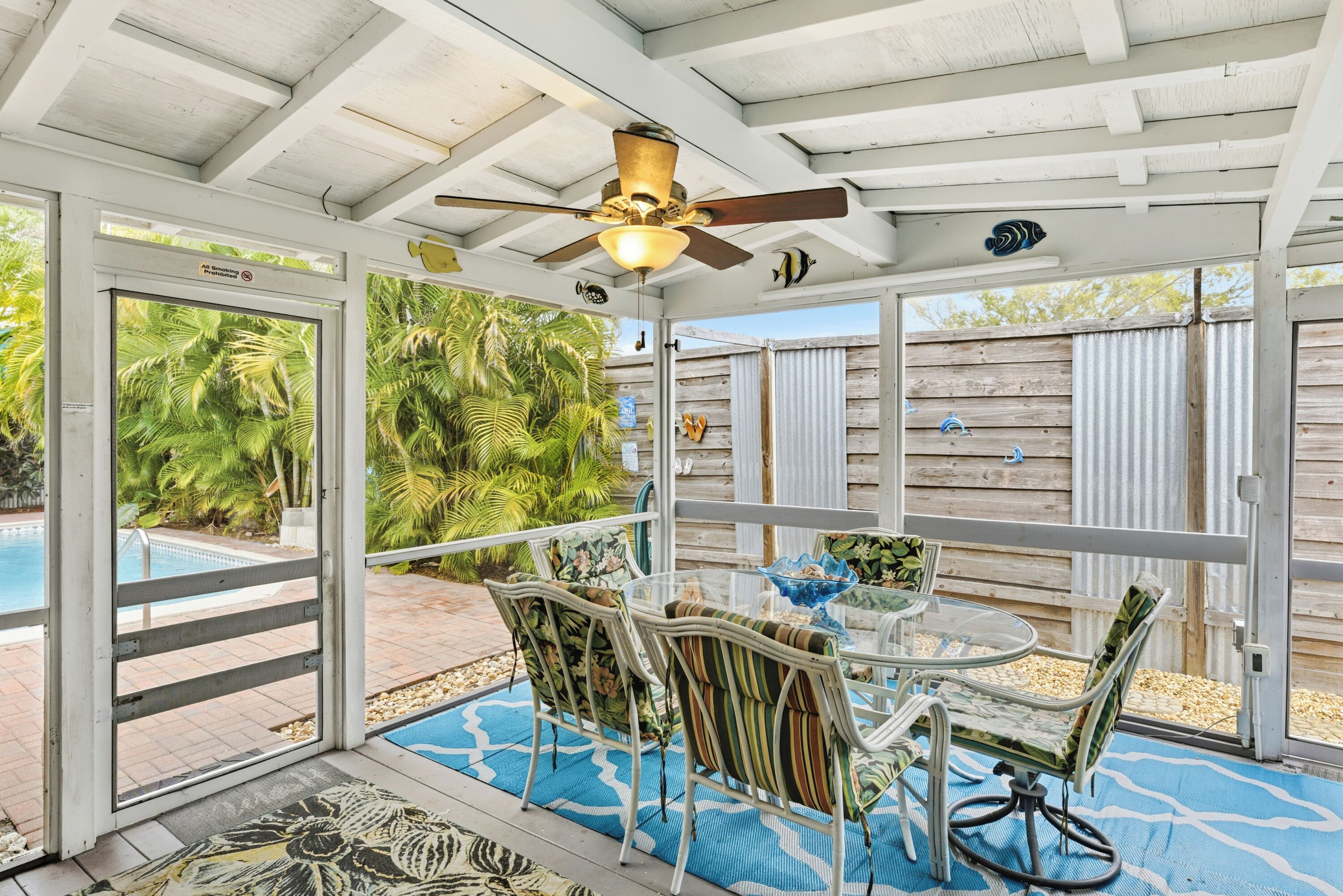 A covered patio area with a glass table and chairs, a ceiling fan, and a view of a pool and palm trees.